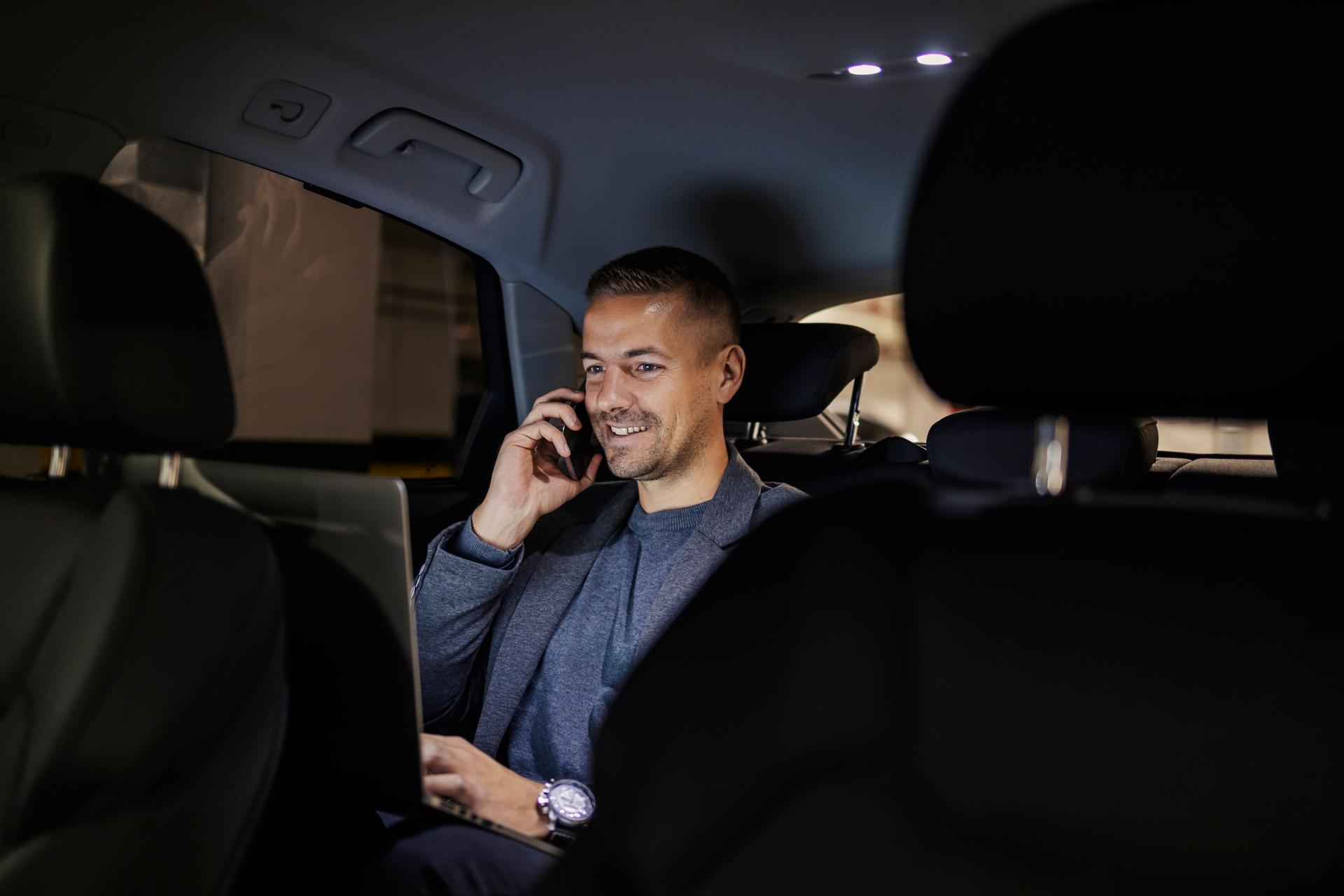 Young smiling businessman on backseat is having phone call and typing on a laptop.